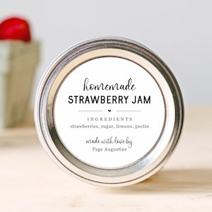 May include: A white jar with a silver lid and a label that reads "homemade STRAWBERRY JAM". The label also lists the ingredients: strawberries, sugar, lemons, and pectin. It says "made with love by Page Augustine".