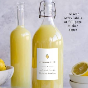 May include: Two clear glass bottles filled with a yellow liquid, labeled "limoncello made with love by Marie and Jonathon". The bottles are sitting on a white surface with lemons in a bowl in the foreground.