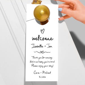 Op de afbeelding: Witte deurhanger met een gouden knop. De tekst luidt "♡ welkom Isabella + Jon Bedankt voor je komst. We zijn zo blij dat je er bent! Geniet van je verblijf! Cara + Michael 3.2.2026"