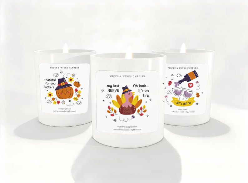 May include: Three white candles with humorous labels. One candle has a pumpkin wearing a witch hat, another has a turkey, and the last has wine glasses. The labels read "thankful for you fuckers", "my last NERVE, It's on fire", and "let's get it".