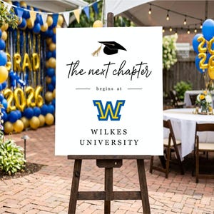 May include: A white sign on a wooden easel with the text "The next chapter begins at Wilkes University." The sign features a graduation cap graphic and the university's logo in blue and yellow. Balloons and decorations are in the background.