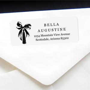 Op de afbeelding: Wit rechthoekig etiket met zwarte tekst en een zwarte strik- en kwastillustratie. Op het etiket staat "BELLA AUGUSTINE" en bevat een adres: 1234 Mountain View Avenue, Scottsdale, Arizona 85302.