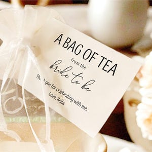 Puede incluir: Una etiqueta blanca con texto negro que dice "A Bag of Tea from the Bride to Be. Thanks you for celebrating with me. Love, Bella." La etiqueta est&aacute; atada a una bolsa de organza blanca.