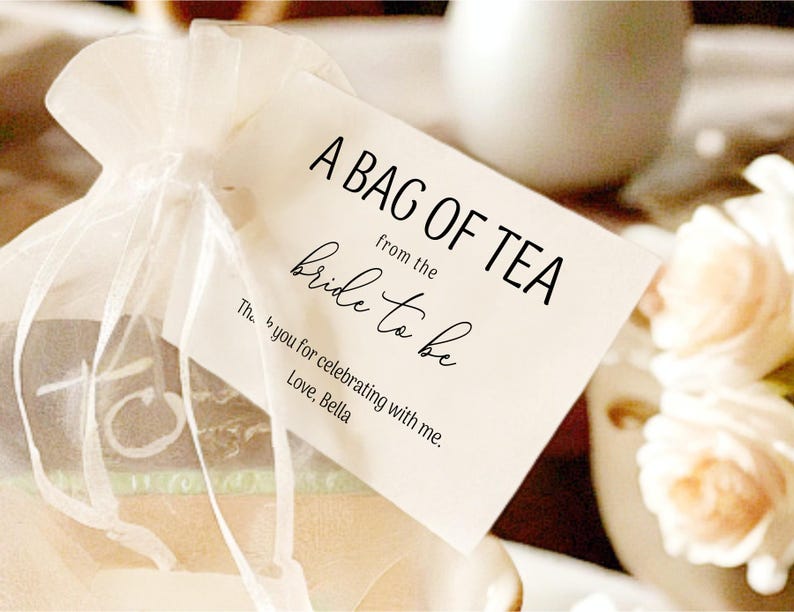 Puede incluir: Una etiqueta blanca con texto negro que dice "A Bag of Tea from the Bride to Be. Thanks you for celebrating with me. Love, Bella." La etiqueta est&aacute; atada a una bolsa de organza blanca.