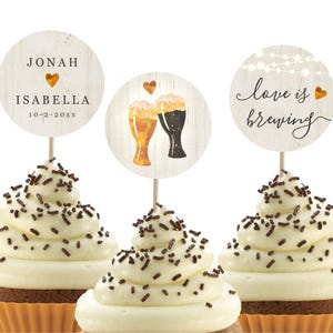 May include: Three cupcakes with white frosting and chocolate sprinkles topped with round paper circles. The circles have text that reads "Jonah Isabella 10-2-20XX", "Love is Brewing" and a design of two beer glasses with hearts.