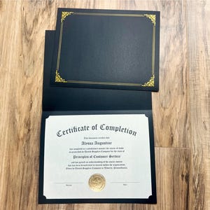 May include: A black certificate holder with gold trim, open to display a certificate. The certificate reads "Certificate of Completion" and includes a gold seal. The holder is on a wooden surface.