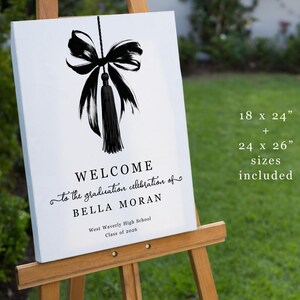 May include: A white graduation welcome sign on an easel. The sign features a black bow and tassel design at the top, with the words "WELCOME" and "to the graduation celebration of BELLA MORAN" in elegant script. The sign is 45.7 x 61 cm and 61 x 66 cm.