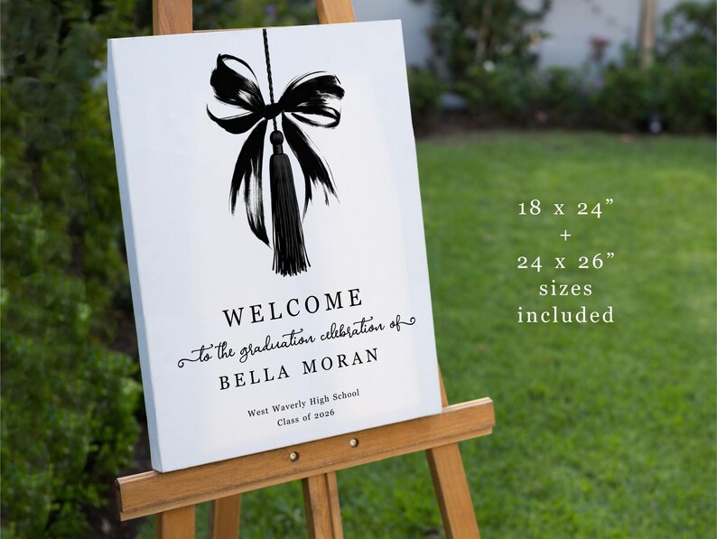 May include: A white graduation welcome sign on an easel. The sign features a black bow and tassel design at the top, with the words "WELCOME" and "to the graduation celebration of BELLA MORAN" in elegant script. The sign is 45.7 x 61 cm and 61 x 66 cm.