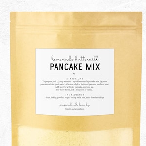 May include: A kraft paper bag with a white label featuring the text "homemade buttermilk PANCAKE MIX." The label includes instructions and ingredients like flour, baking powder, sugar, and mini chocolate chips. The text "prepared with love by Marie and Jonathan" is also visible.