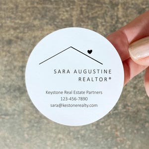 Pu&ograve; includere: Un biglietto da visita rotondo e bianco con il testo "SARA AUGUSTINE REALTOR" e le informazioni di contatto. Il biglietto ha un design minimalista con un contorno di casa e un simbolo a forma di cuore. Il biglietto &egrave; tenuto in mano.