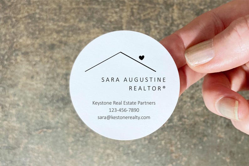 Pu&ograve; includere: Un biglietto da visita rotondo e bianco con il testo "SARA AUGUSTINE REALTOR" e le informazioni di contatto. Il biglietto ha un design minimalista con un contorno di casa e un simbolo a forma di cuore. Il biglietto &egrave; tenuto in mano.
