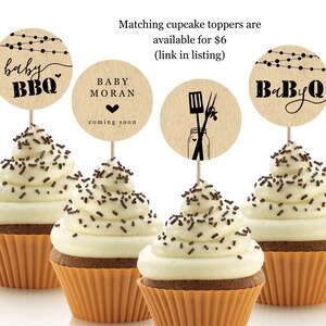 May include: Four cupcakes with white frosting and chocolate sprinkles topped with round brown paper cupcake toppers. The toppers have black text that says "baby BBQ", "BABY MORAN coming soon", and "BaByQ". One topper has a black illustration of a jar with a spatula and tongs.