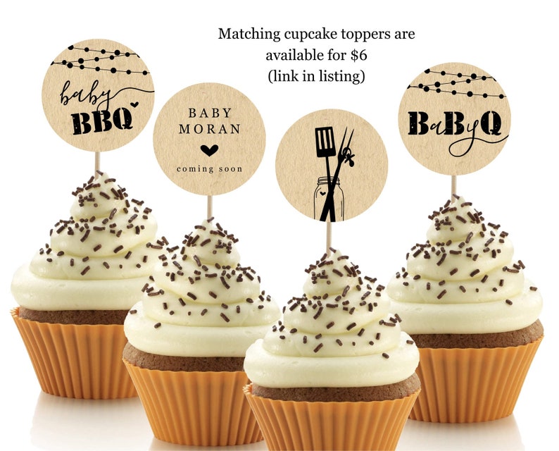 May include: Four cupcakes with white frosting and chocolate sprinkles topped with round brown paper cupcake toppers. The toppers have black text that says "baby BBQ", "BABY MORAN coming soon", and "BaByQ". One topper has a black illustration of a jar with a spatula and tongs.