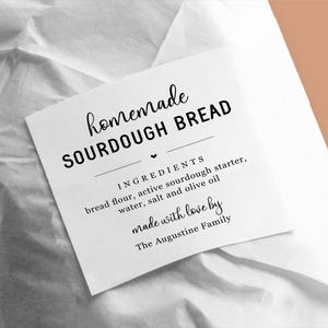 May include: A white paper bag with a label that reads "homemade sourdough bread". The label lists the ingredients: bread flour, active sourdough starter, water, salt and olive oil. It also says "made with love by The Augustine Family."