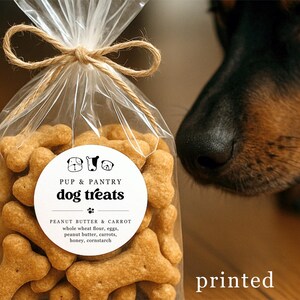 May include: A clear cellophane bag tied with twine, filled with bone-shaped dog treats. The label reads "Pup & Pantry dog treats" with peanut butter and carrot ingredients. A dog's nose is visible in the background.