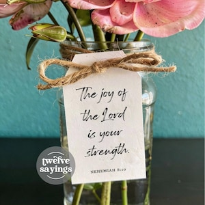 May include: A clear glass jar filled with pink flowers, featuring a white rectangular paper tag tied with brown twine. The tag displays the black printed text: The joy of the Lord is your strength. NEHEMIAH 8:10. A circular logo reads twelve sayings.