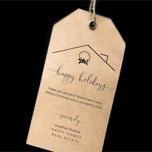 Puede incluir: Una etiqueta de regalo beige con un contorno de casa negro y las palabras "happy holidays" en cursiva. La etiqueta incluye un saludo festivo y el nombre "Jonathon Thomas" con "HAPPY HOMES REAL ESTATE" debajo.