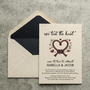 May include: A cream-coloured wedding invitation card and matching envelope with a black interior. The card features a brown rope tied into a heart shape, red hearts, and black text reading "we tied the knot" and "ISABELLA & JACOB" with event details.