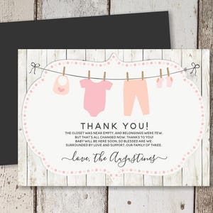 Op de afbeelding: Roze babykleding hangt aan een waslijn tegen een witte achtergrond met de tekst "Bedankt! De kast was bijna leeg en er waren maar weinig spullen. Maar dat is nu veranderd, dankzij jou! De baby is binnenkort hier, we zijn zo gezegend. Omringd door liefde en steun, ons gezin van drie. Met liefde, de Augustines."