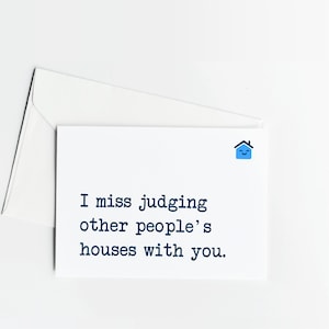 以下が含まれることがあります： 「I miss judging other people's houses with you.」という濃い青色のテキストと、カードの右上隅に配置された小さな水色の笑顔の家のアイコンが特徴の、白いグリーティングカードと封筒のセット。