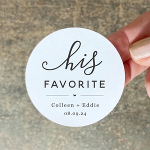 Op de afbeelding: Ronde witte sticker met de tekst "his favorite" in zwarte cursieve letters. Onder de tekst staat een klein hartje en de namen "Colleen + Eddie" en de datum "08.09.24" in zwart.