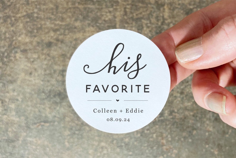 Op de afbeelding: Ronde witte sticker met de tekst "his favorite" in zwarte cursieve letters. Onder de tekst staat een klein hartje en de namen "Colleen + Eddie" en de datum "08.09.24" in zwart.