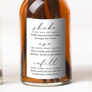 Op de afbeelding: Een doorzichtige glazen fles gevuld met donkere vloeistof en vanillestokjes, voorzien van een wit rechthoekig etiket met zwarte tekst: shake every now and again, use the little black specks en refill with vodka once empty.