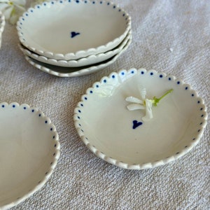 Peut inclure: Ensemble de quatre plats en céramique blanche avec des bords festonnés et un motif de points bleus. Chaque plat a un petit cœur bleu au centre.
