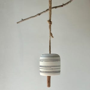 May include: A white ceramic bell with black horizontal stripes, suspended by a natural fibre rope. The bell has a wooden clapper and is hanging from a small tree branch. The design is minimalist and rustic.