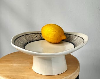 Puede incluir: Un cuenco de cerámica blanca sobre pedestal con una base ancha y poco profunda que presenta un patrón de líneas negras a lo largo del borde interior. Un limón amarillo brillante descansa en el centro del cuenco, con acabado liso y brillante.