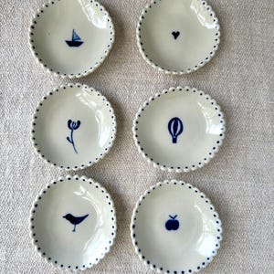 Peut inclure: Six petites assiettes en céramique blanche avec des bords bleus festonnés et des motifs bleus. Les motifs comprennent un voilier, un cœur, une fleur, une montgolfière, un oiseau et une pomme.