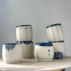 May include: Six small, off-white ceramic cups with dark blue rims and blue floral and linear patterns. The cups are stacked on a light-coloured wooden surface. The background is plain white.