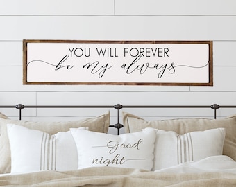 May include: A white bedroom with a wooden headboard and a sign above the bed that reads "You will forever be my always". There are two nightstands with lamps and plants on them.