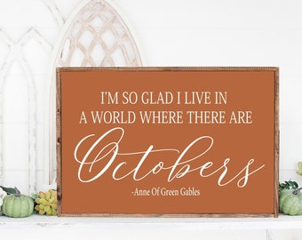 May include: A brown wooden sign with white text that reads "I'm so glad I live in a world where there are Octobers - Anne of Green Gables". The sign is sitting on a white mantel with a garland of green leaves and small pumpkins.