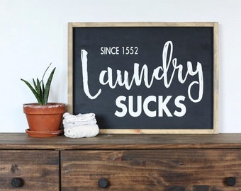 May include: A dark brown wooden dresser with four drawers and black knobs. A black framed sign with white text reads "Since 1552 Laundry Sucks". A potted succulent plant and a stack of white cloth are on top of the dresser.