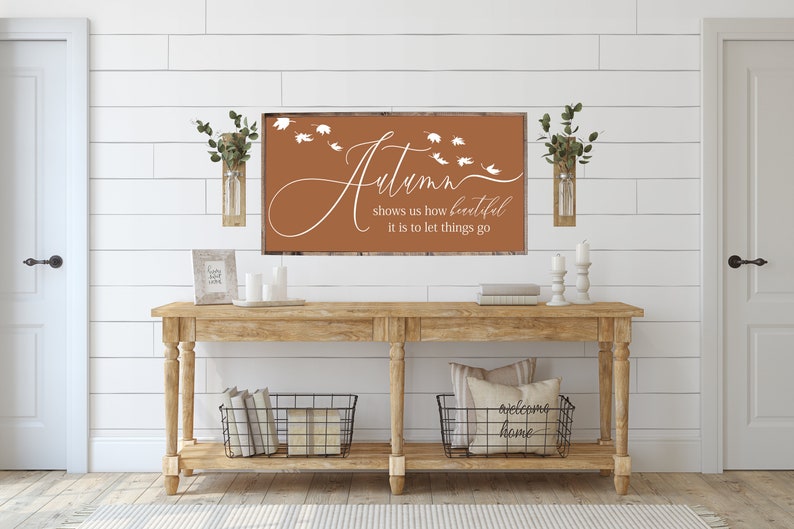 May include: A brown wooden sign with white text that reads "Autumn shows us how beautiful it is to let things go". The sign is hanging on a white wall with white shiplap panels. There is a wooden console table with two black wire baskets underneath. The table is decorated with candles, greenery, and a pillow that reads "welcome home".