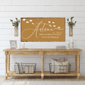 May include: A wooden sign with a mustard yellow background and white text that reads "Autumn shows us how beautiful it is to let things go". The sign is hanging on a white shiplap wall above a wooden console table with two black wire baskets. There are two small glass vases with greenery on either side of the sign.