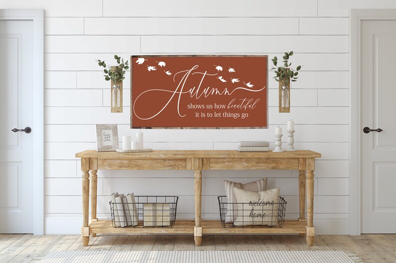 May include: A rustic wooden console table with two black wire baskets, a white pillow with the text "welcome home", and a framed sign with the text "Autumn shows us how beautiful it is to let things go" on a white shiplap wall.