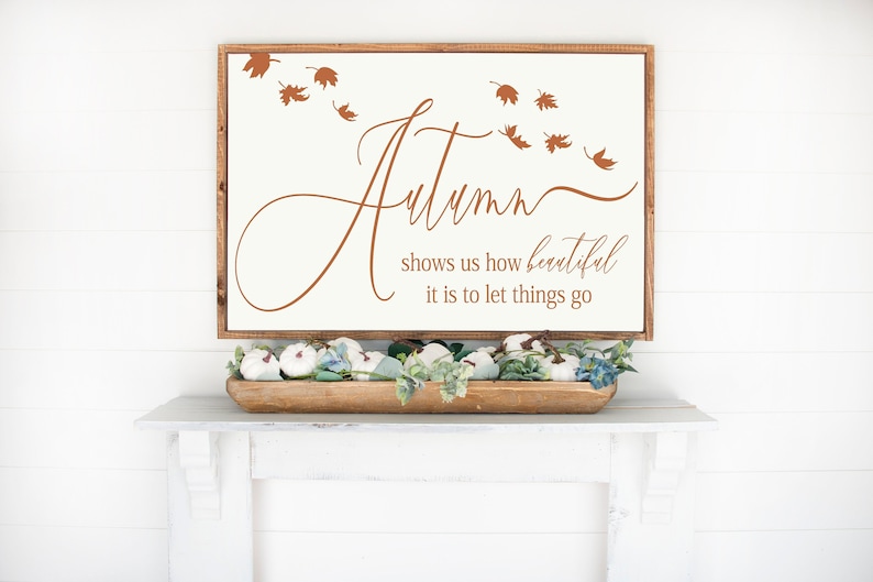 May include: A white wooden sign with brown text that reads "Autumn shows us how beautiful it is to let things go". There are brown leaf silhouettes scattered around the text. The sign is sitting on a white shelf with a wooden bowl filled with white pumpkins and greenery.