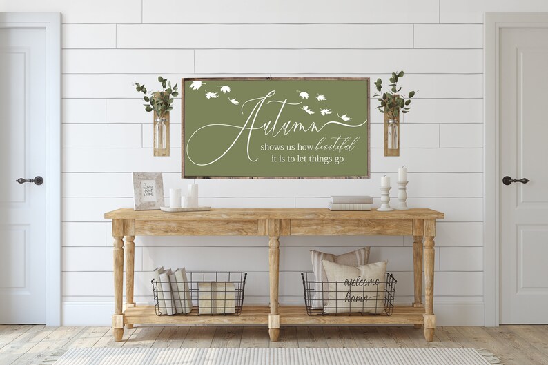 May include: A wooden sign with a green background and white text that reads "Autumn shows us how beautiful it is to let things go". The sign is hanging on a white wall above a wooden console table with two black wire baskets and a framed print.