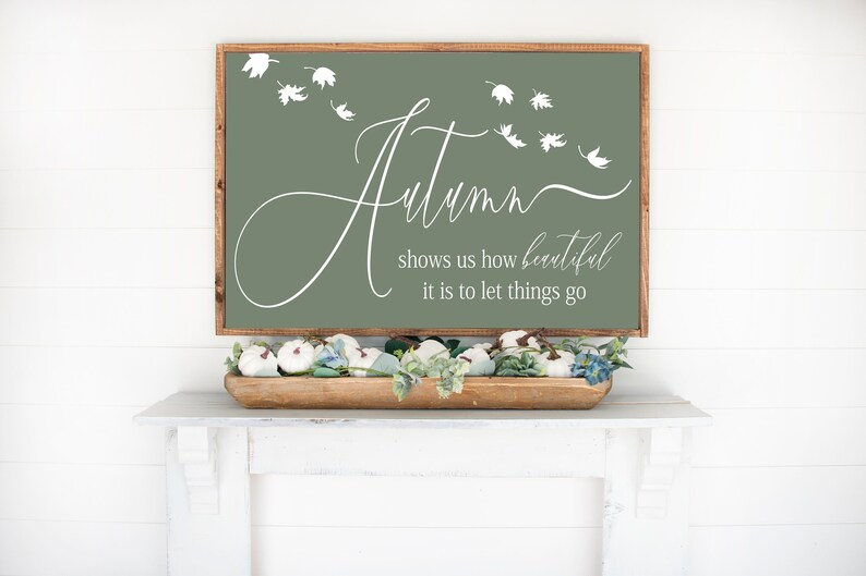 May include: A sage green wooden sign with white lettering that reads "Autumn shows us how beautiful it is to let things go". The sign is decorated with white leaf cutouts.