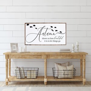 May include: A wooden sign with the text "Autumn shows us how beautiful it is to let things go" in black script. The sign is hanging on a white shiplap wall above a wooden console table with two black wire baskets and candles.