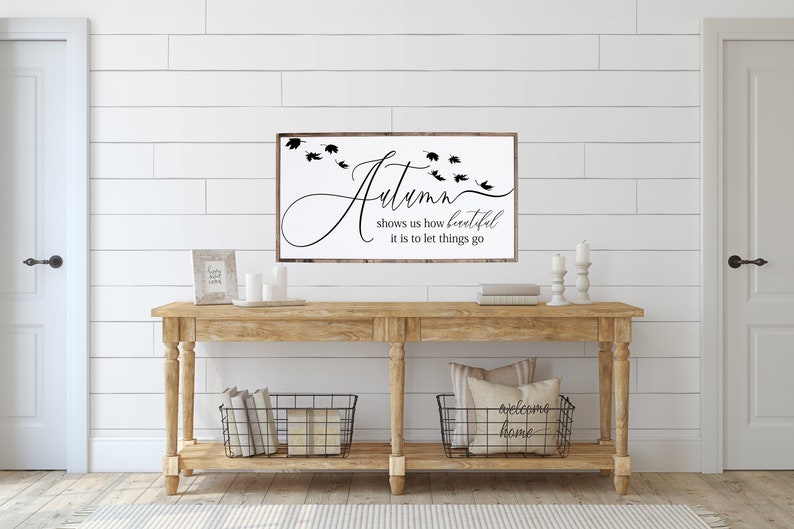 May include: A wooden sign with the text "Autumn shows us how beautiful it is to let things go" in black script. The sign is hanging on a white shiplap wall above a wooden console table with two black wire baskets and candles.