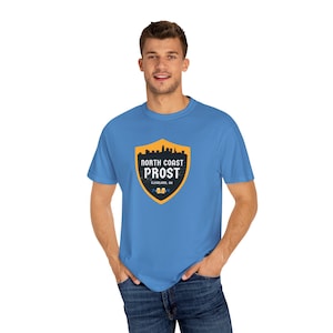May include: A light blue t-shirt featuring a shield-shaped graphic with the words "NORTH COAST PROST" and "CLEVELAND, OH" in white and gold. The graphic includes a skyline silhouette and two beer mugs.