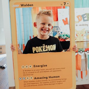 May include: A yellow and orange cardboard cutout of a Pokemon card with a boy holding it in front of his face. The card says "Walden is 7" and has a picture of a Pokemon character on it. The card also has text that says "Energize" and "Amazing Human".