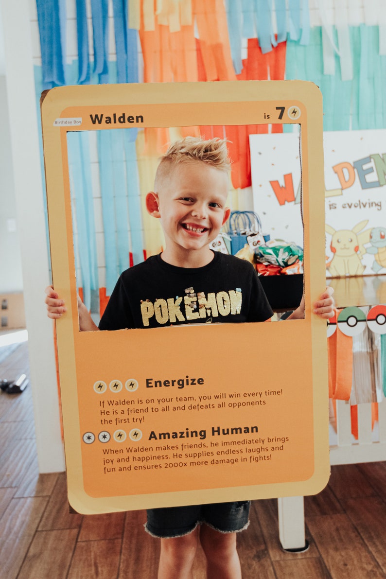 May include: A yellow and orange cardboard cutout of a Pokemon card with a boy holding it in front of his face. The card says "Walden is 7" and has a picture of a Pokemon character on it. The card also has text that says "Energize" and "Amazing Human".