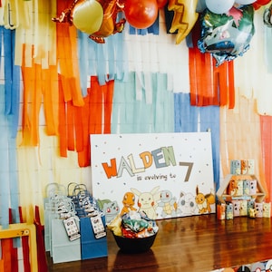 May include: A Pokemon-themed birthday party decoration with a sign that says "Walden is evolving to 7". The sign is surrounded by colourful balloons and streamers. There are also small Pokemon figurines and gift bags.