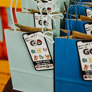 May include: Blue and light blue paper gift bags with handles. Each bag has a tag attached with the text "We're sad you have to go" and a Pokemon character graphic. The tags also include the text "Thanks for celebrating Walden evolving to 7!" and a row of Pokemon symbols.