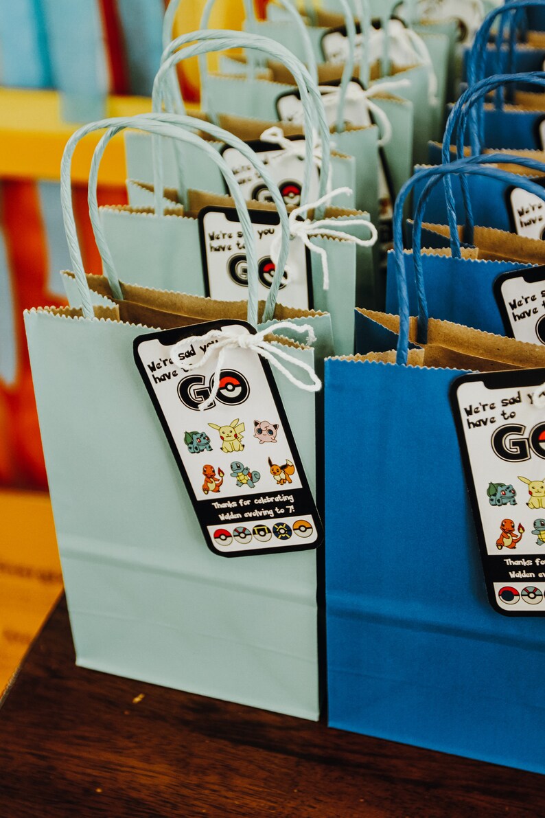 May include: Blue and light blue paper gift bags with handles. Each bag has a tag attached with the text "We're sad you have to go" and a Pokemon character graphic. The tags also include the text "Thanks for celebrating Walden evolving to 7!" and a row of Pokemon symbols.