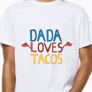 Puede incluir: Camiseta blanca con el texto "DADA LOVES TACOS" en letras de colores. El texto est&aacute; rodeado de dos paraguas rojos.